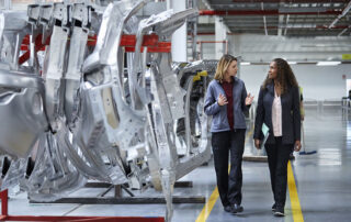 Female engineers discussing by car chassis. Full length of professionals are walking on aisle in factory. Colleagues are in automobile industry.
