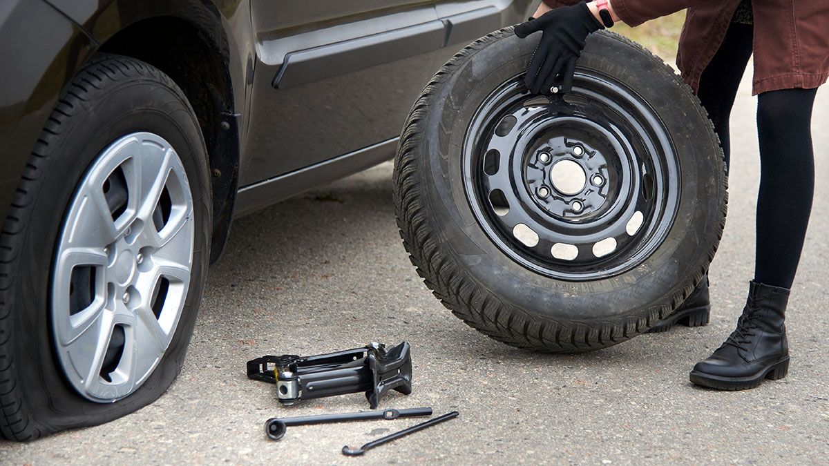 A young woman rolls spare tire near her car with a flat tire, trouble on the road.