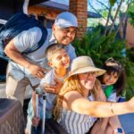 Family taking a selfie while packing their car for a summer road trip, with luggage nearby and The General Insurance logo in the corner.