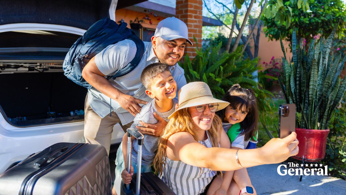 Family taking a selfie while packing their car for a summer road trip, with luggage nearby and The General Insurance logo in the corner.