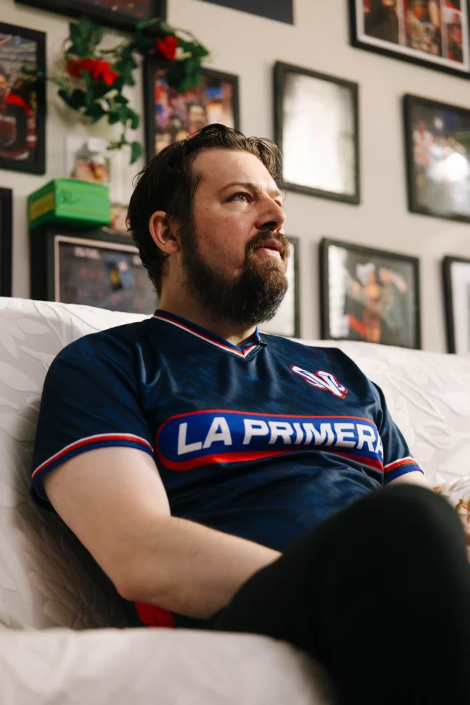 A WWE Fan of the Year finalist wearing a blue soccer jersey sits on a couch, looking off-camera with a focused expression in a room decorated with framed sports photos.
