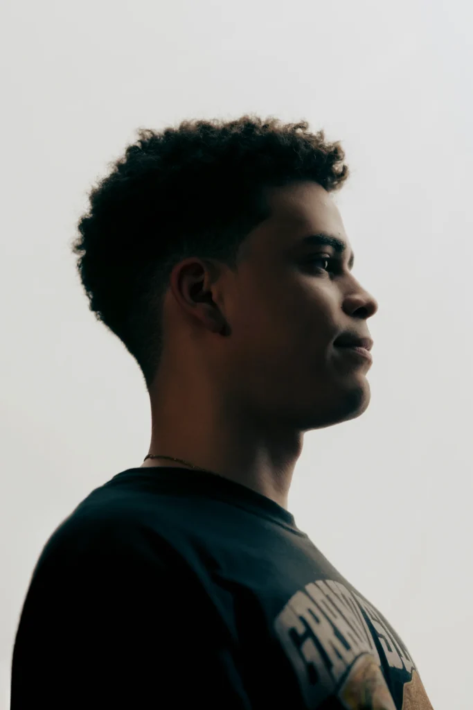 A side-profile portrait of a WWE Fan of the Year finalist wearing a black graphic T-shirt, looking ahead with a calm, determined expression.