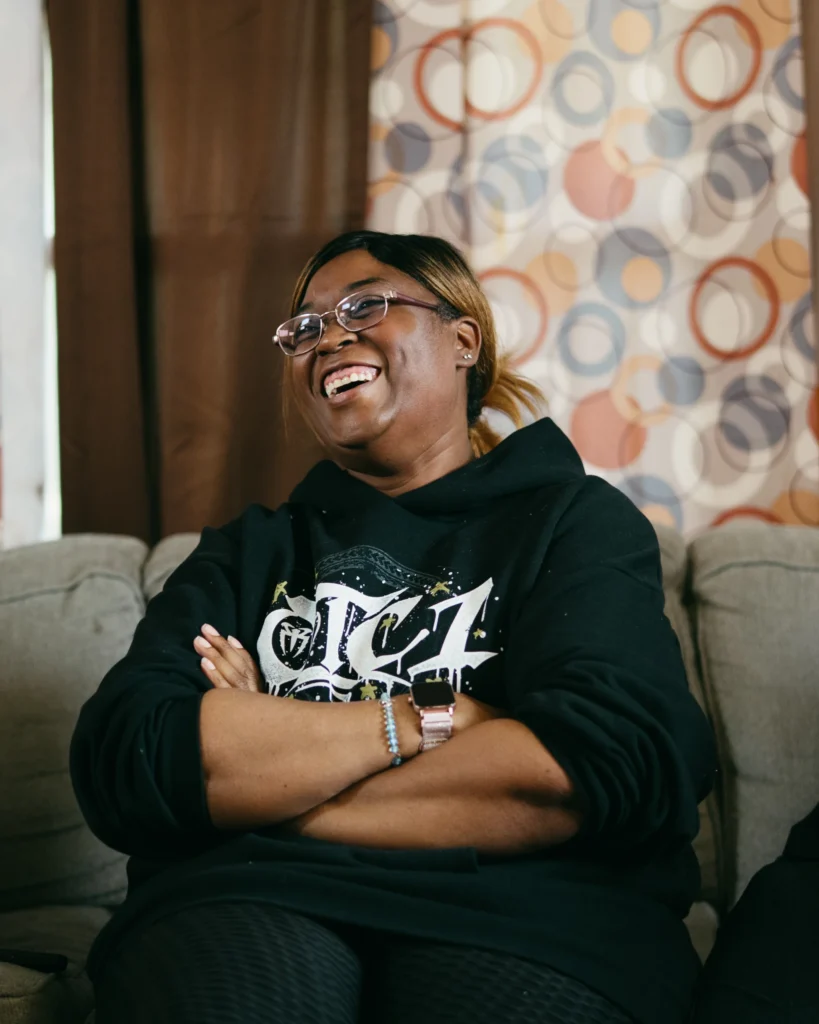 A smiling WWE Fan of the Year finalist wearing a black WWE hoodie sits on a couch with arms crossed, laughing during an interview.